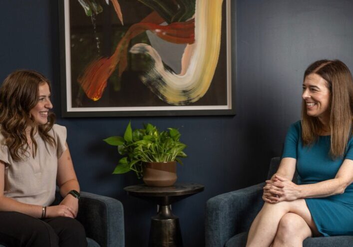 Dr. Amy Fogelman, MD, in a teal dress, sits across from a smiling client in a professional office setting with navy walls, abstract artwork, and a green plant between them. This image reflects High Rock Experts’ one-on-one coaching for medical professionals and expert witness consulting services that support both attorneys and medical professionals nationwide.