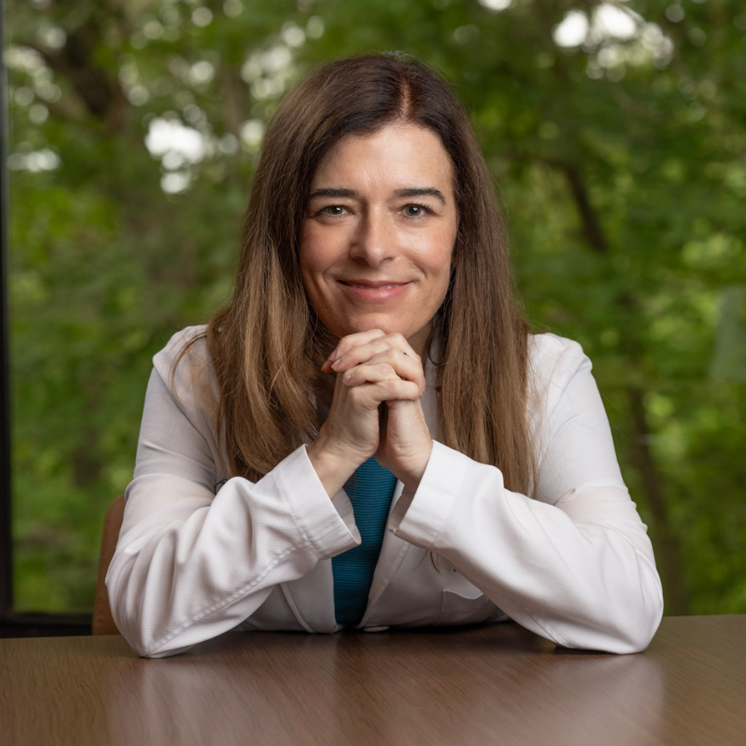 Amy Fogelman, MD, founder of High Rock Experts, sits at a desk in a white coat with hands clasped and smiling. She is a physician consultant who trains medical professionals to serve as expert witnesses and connects them with attorneys for legal case review.