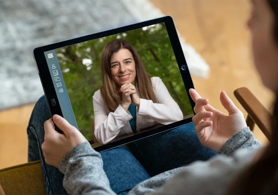 A person participates in a virtual consultation on a tablet with Dr. Amy Fogelman, MD, who is wearing a white coat and smiling warmly. Dr. Fogelman is a leading medical expert witness and founder of High Rock Experts, offering expert witness training and physician case reviews. This image represents remote expert witness consulting services for attorneys nationwide.